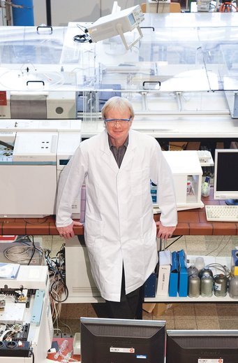 Portrait of Ferdi Schüth. Schüth is wearing a lab coat and protective goggles, leaning against a laboratory counter with analytical equipment whose shape and size are reminiscent of printers. In the background, there is a flat box consisting of acrylic glass on a table, filling the entire width of the picture.